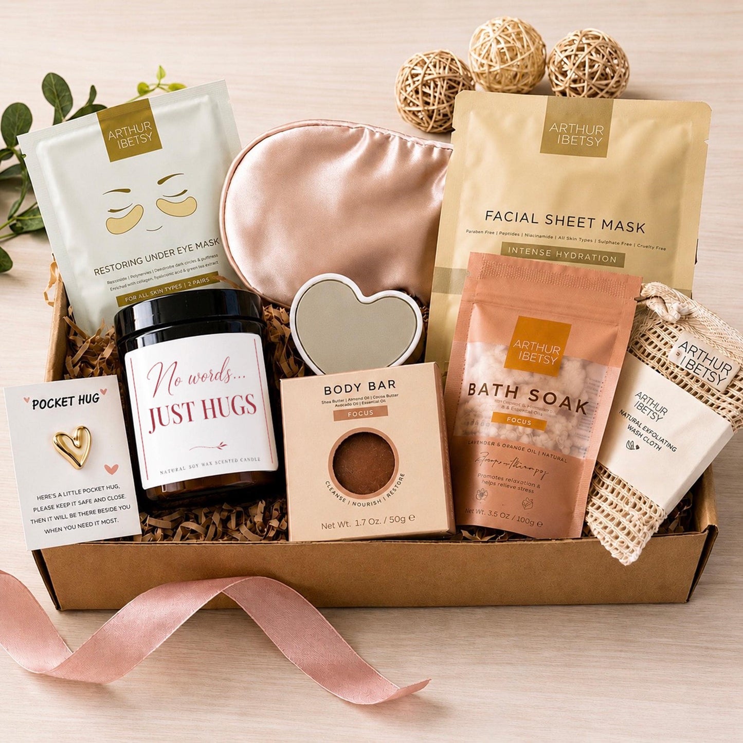 A self-care gift set in a cardboard box, featuring a pink satin eye mask, a candle with the text "No words... JUST HUGS", a body bar, bath soak, and an under-eye mask. The box is tied with a pink ribbon.