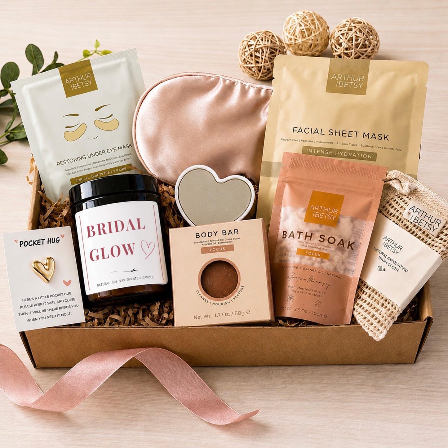 A self-care gift set in a cardboard box. It contains a candle, eye mask, body bar, bath soak, and under-eye masks. The candle is labelled "Bridal Glow". The box is filled with paper and tied with a pink ribbon.