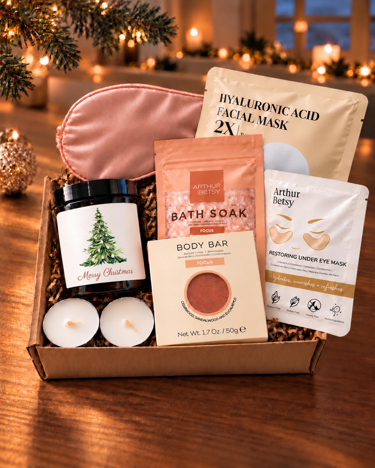 Christmas spa gift box with an amber candle, pink satin sleep mask, bath soak, soap, facial mask, under-eye mask and two tealights, styled on a wooden table.